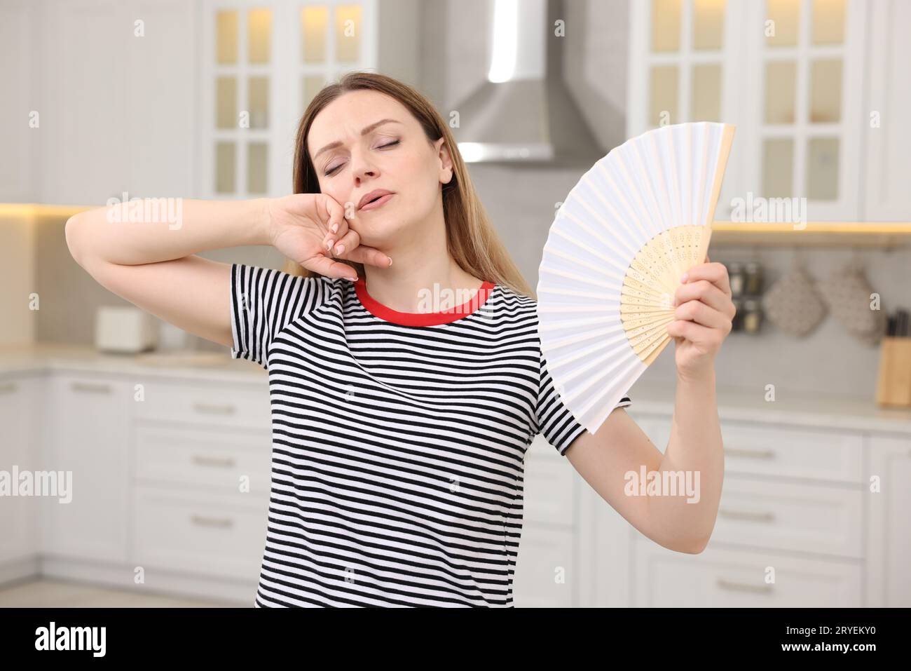 Woman with hand fan suffering from heat in kitchen Stock Photo
