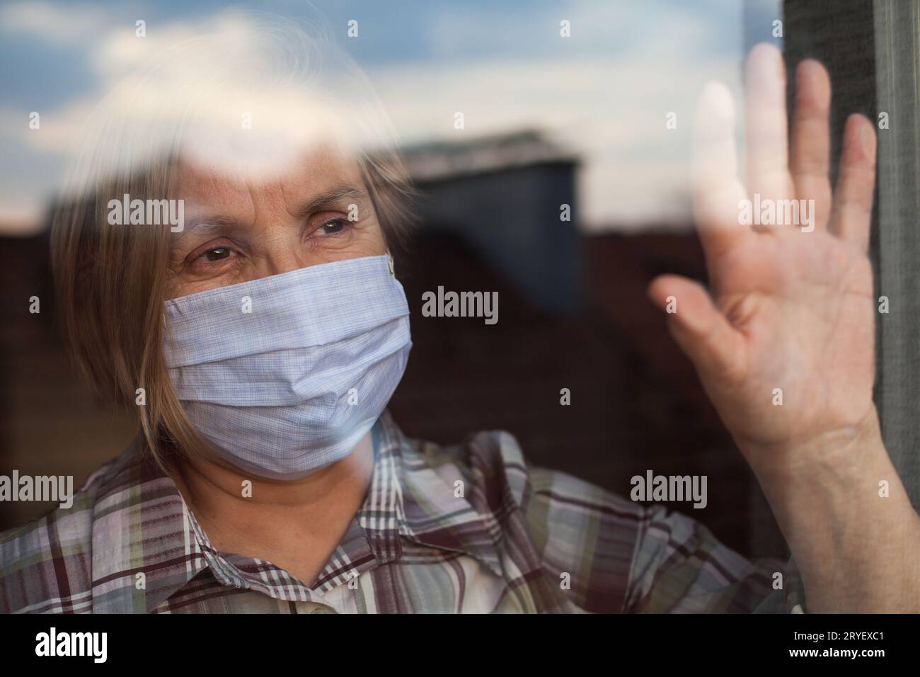 Portrait of elderly senior citizen wearing face mask looking through room window Stock Photo
