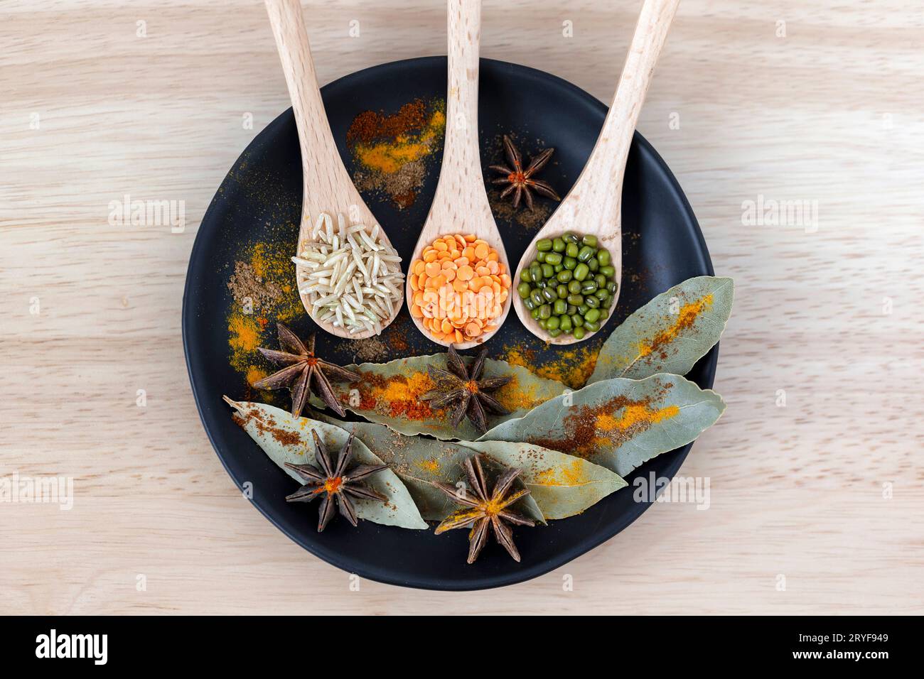 Rice, moong beans and dal in wooden spoons next to star anise seeds, bay leaf and colorful