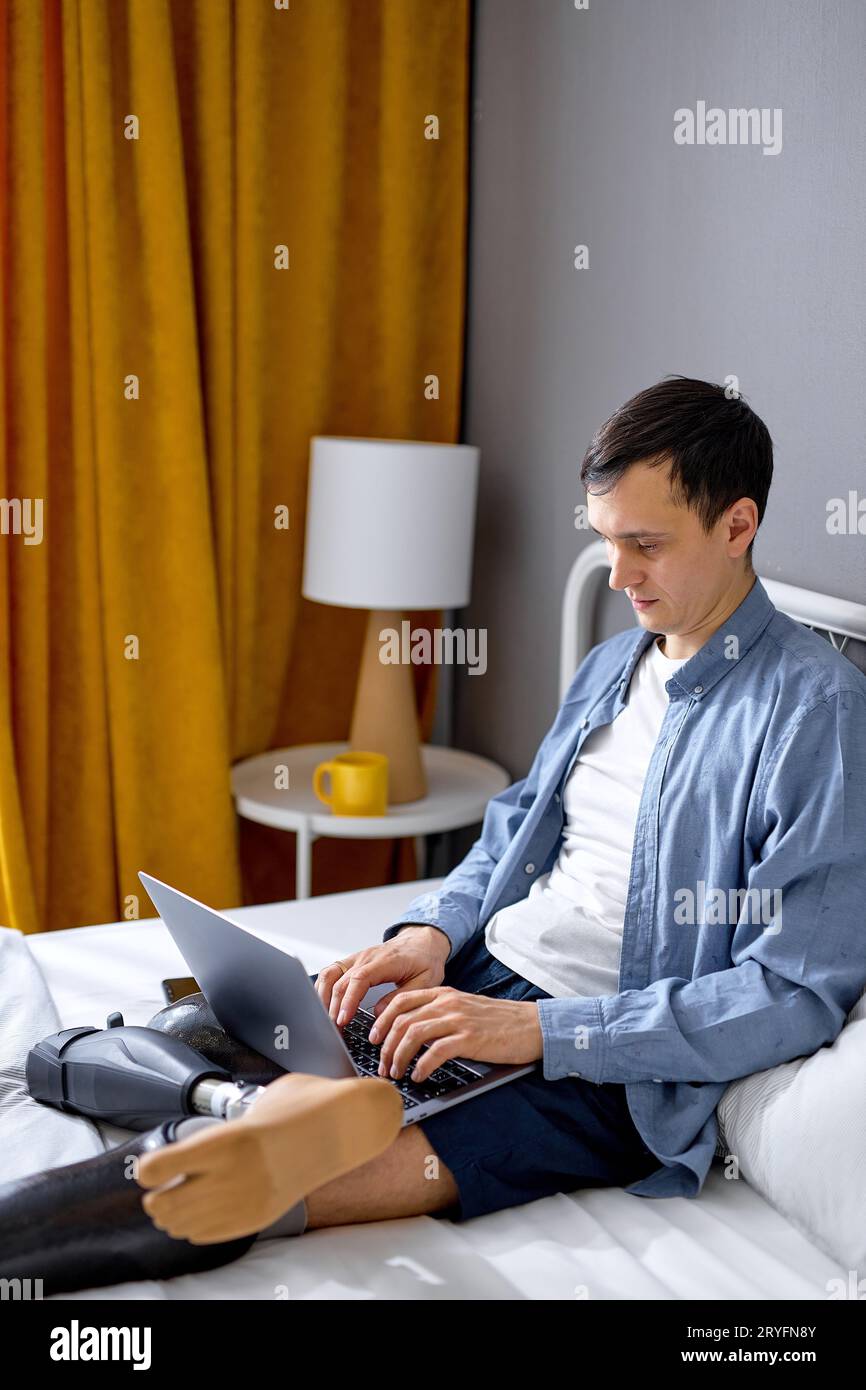 concentrated young man with prosthesis on legs using laptop for remote job at home, sitting on bed. Confident Caucasian guy with physical disability h Stock Photo