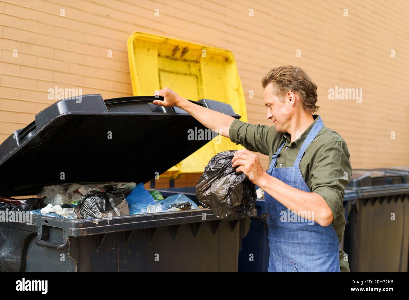 Caucasian handsome man employee open can full of garbage to throw plastic bag with trash wearing apron. Wrong unsorted garbage c Stock Photo