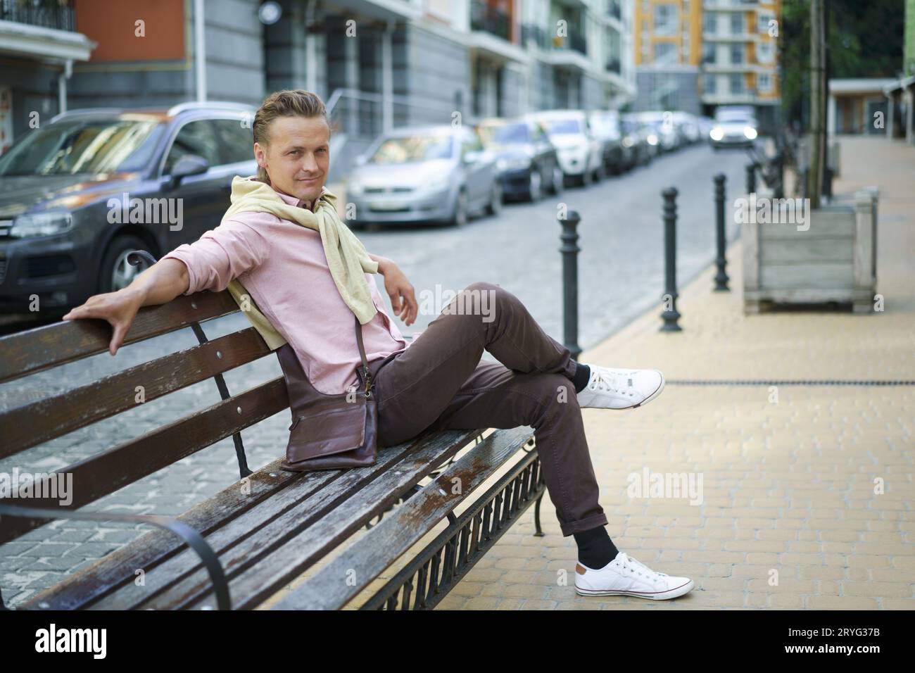 Handsome man sits on the bench with legs crossed spend time in urban city near his office or home, wearing pink shirt and should Stock Photo