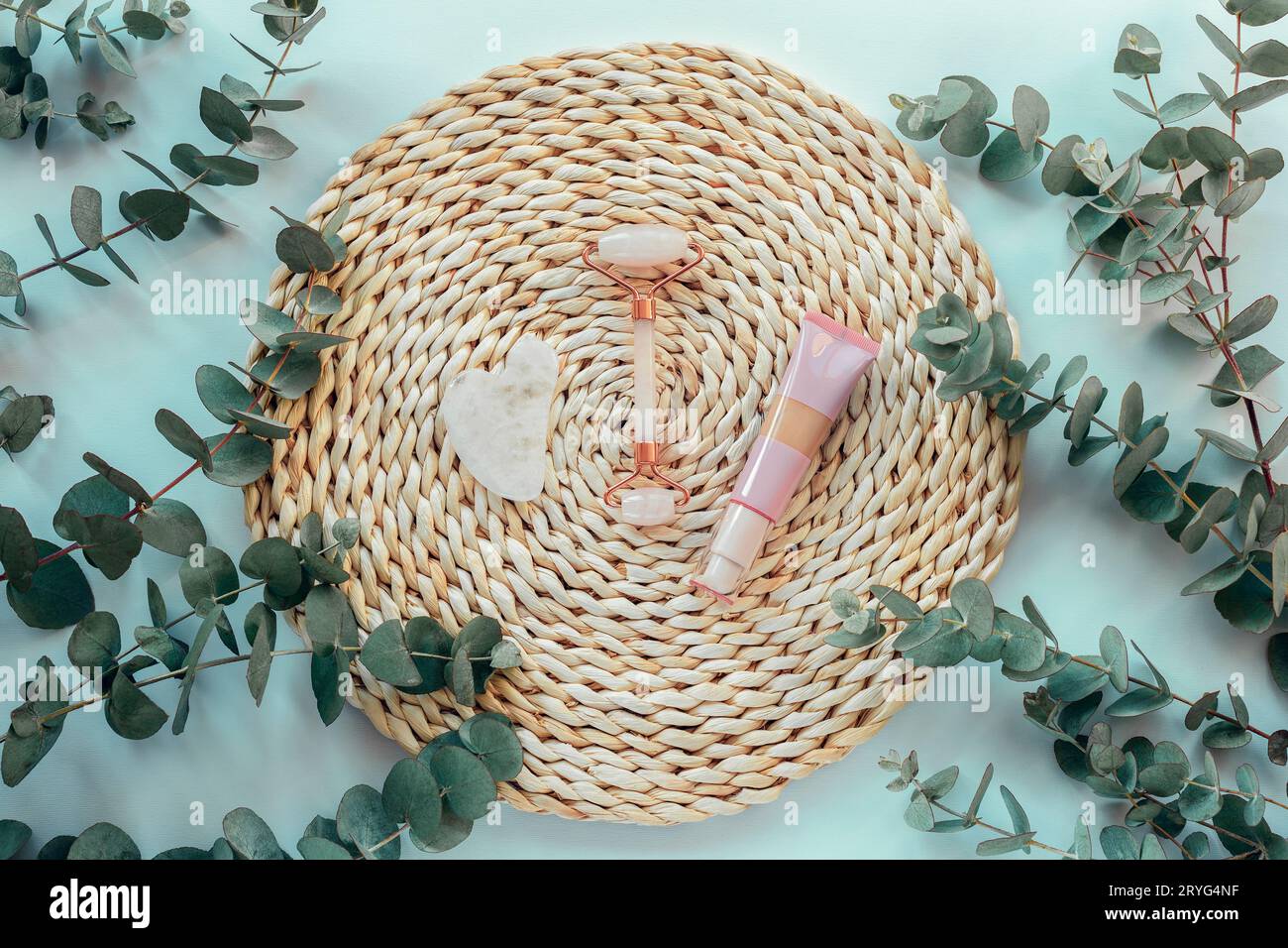 Gua sha stone, face roller and cream tube on a wicker place mat with eucalyptus branches on light blue table. Skin care, beauty treatment concept. Top Stock Photo