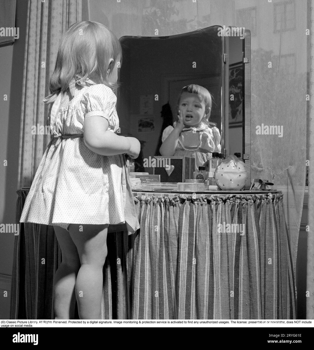 In the 1950s. A little girl is standing in front of her mothers makeup table and mirror and puts on some kind of beauty cream on her face, but does not look so happy. Sweden 1951 Conard ref 1708 Stock Photo