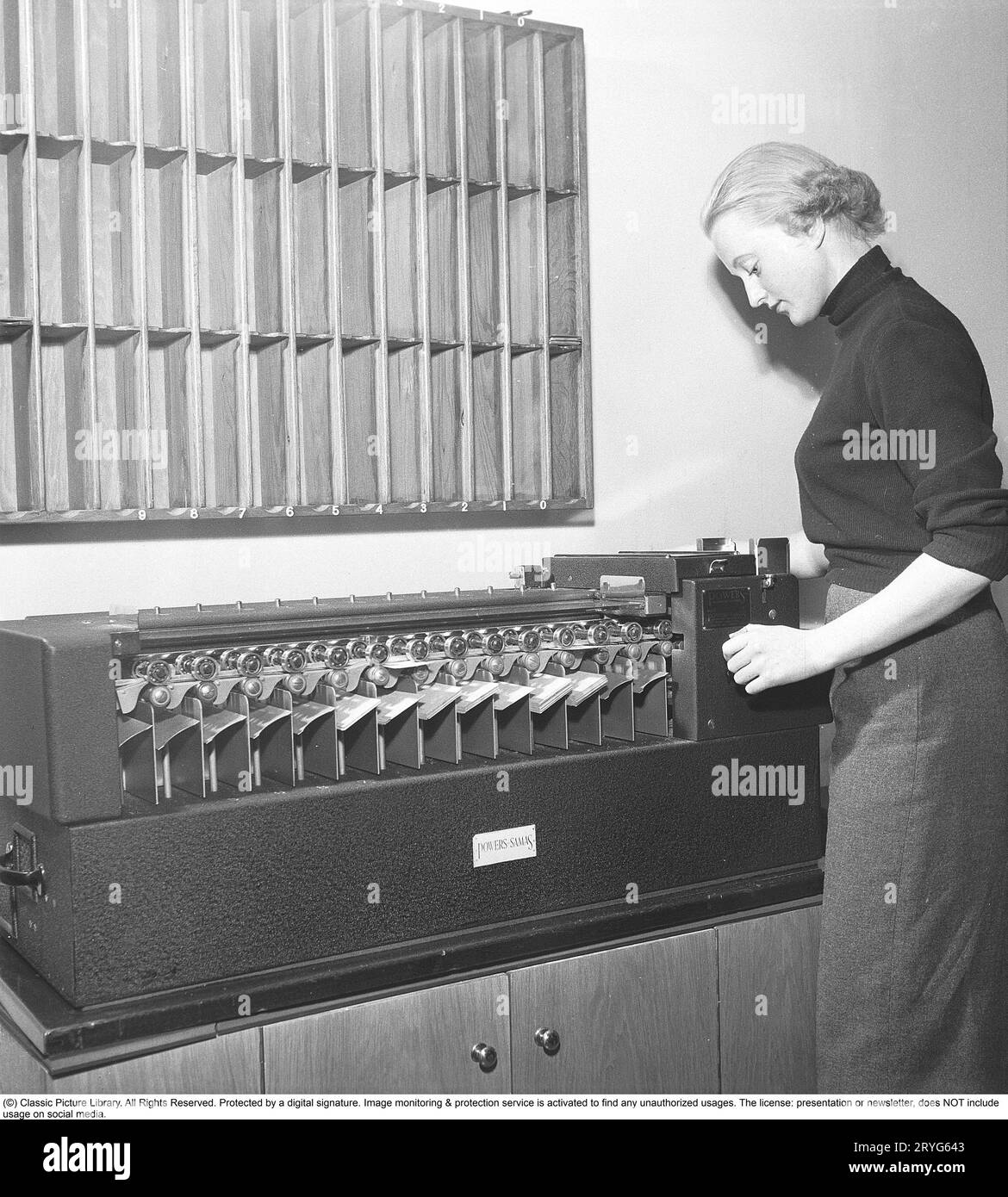 In the 1950s. A female clerk at punch card reader machine overlooking it's function and the feed of  the punch cards into the machine. Punched cards was created by another machine, entering information, creating the punch cards as a storage medium where the punched holes in the paper card were the input information. The picture shows the reader sorting the cards after having read the information in the punch holes. The machine is made by the british manufacturer Powers-Samas who developed the encryption machine, far better and safer then the german Enigma.  Sweden 1954. Kristoffersson ref BM85 Stock Photo