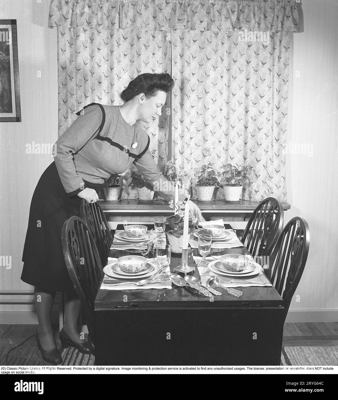 In the 1940s. A woman has set a table with plates and glasses and is seen attending to the details like the napkins. Perhaps friends are coming over for dinner. Sweden 1945. Kristoffersson ref M121-3 Stock Photo