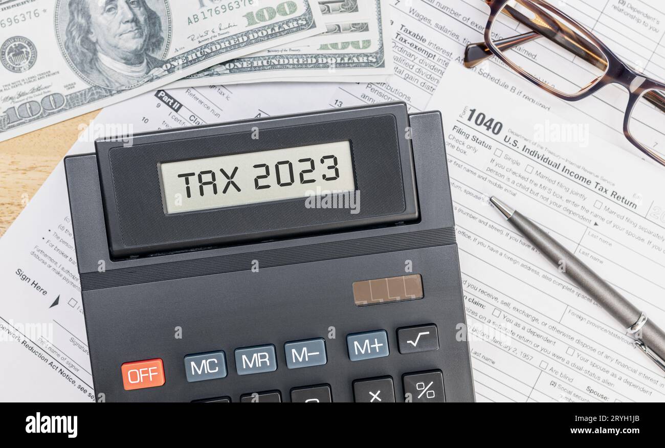 Calculator with TAX 2023 text on screen, 1040 form, dollar banknotes and eyeglasses on desk. Top view Stock Photo