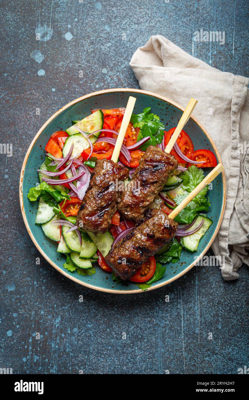 Grilled skewer meat beef kebabs on sticks served with fresh vegetables salad on plate on rustic concrete background from above. Stock Photo