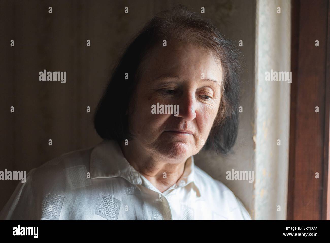 Elderly woman standing by the window, looking sad Stock Photo