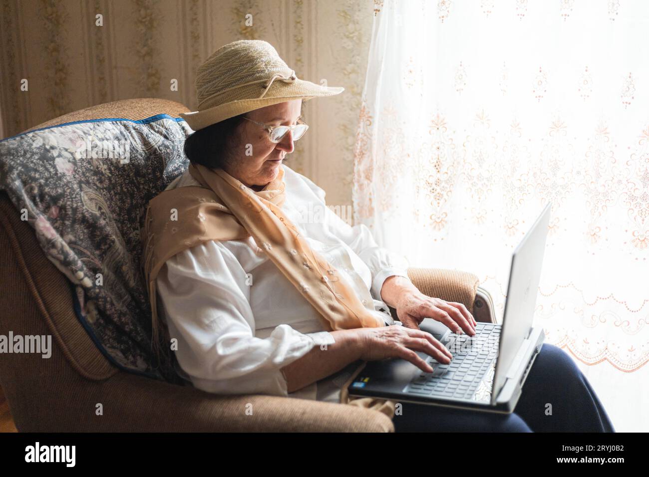 Elderly woman working on a laptop, sitting in a chair by the window Stock Photo
