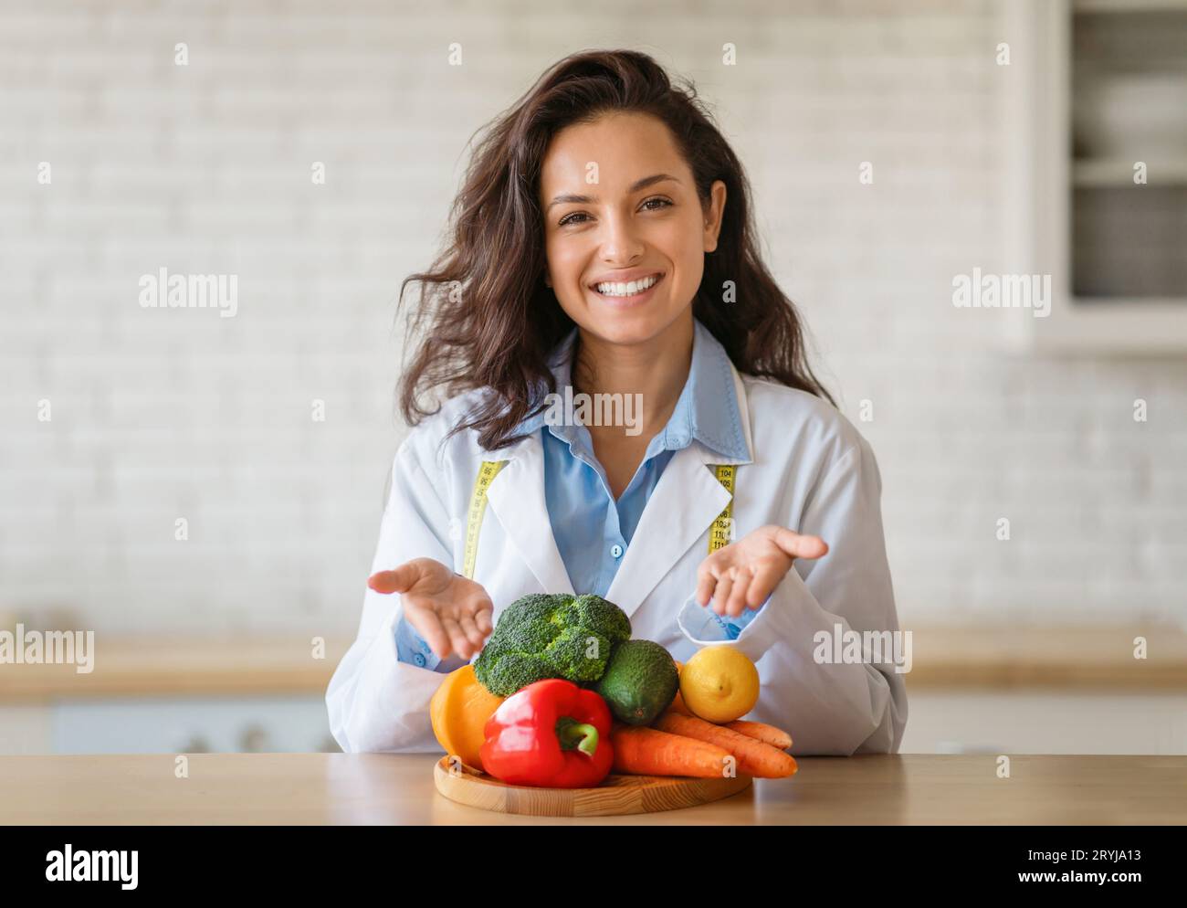 Portrait of cheerful dietitian in lab coat pointing at fresh fruits and vegetables on table, smiling at camera at clinic Stock Photo