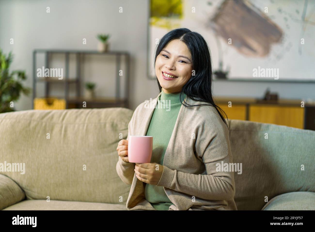 Mature Asian woman enjoys hot cup of tea while relaxing on her cozy living room sofa Stock Photo