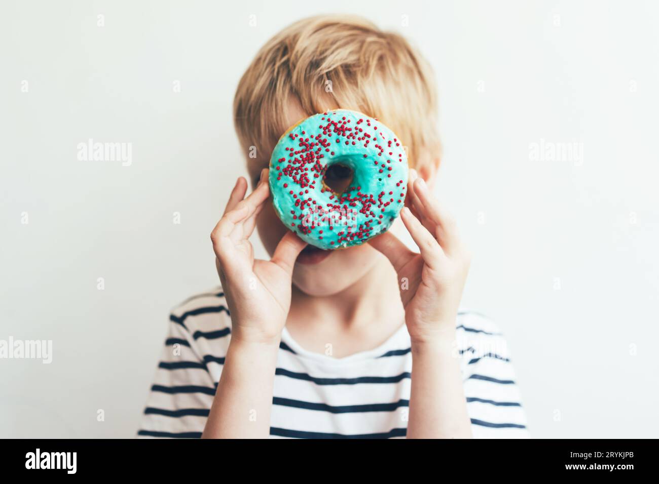 A blond boy looks through a donut in blue glaze. Portrait of a child with pastries close-up. Stock Photo