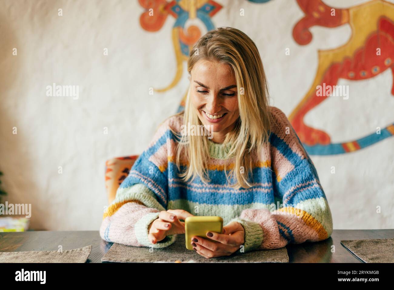 A young woman in a striped colored sweater with a mobile phone at a table in a cafe laughs and watches social media. Stock Photo