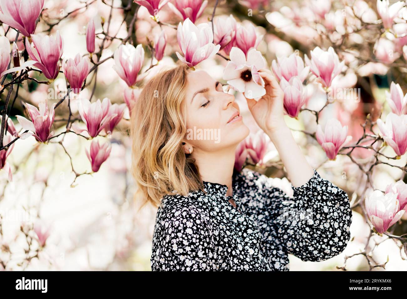 Young attractive woman in the park near a blooming magnolia tree inhales the aroma of flowers and enjoys the spring. Stock Photo