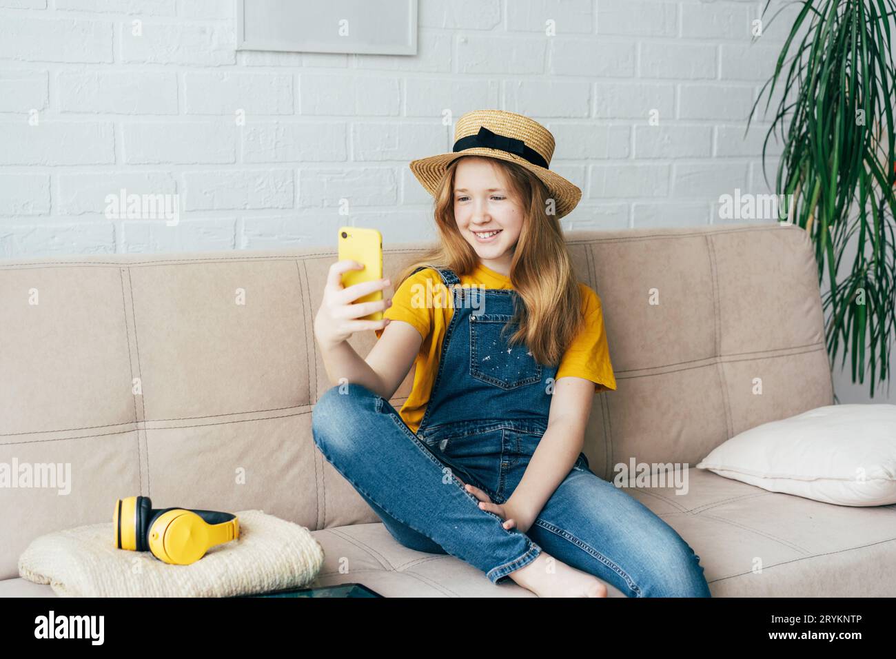 Cheerful redhead girl in a straw hat and denim overalls posing and taking a selfie on a smartphone. Stock Photo