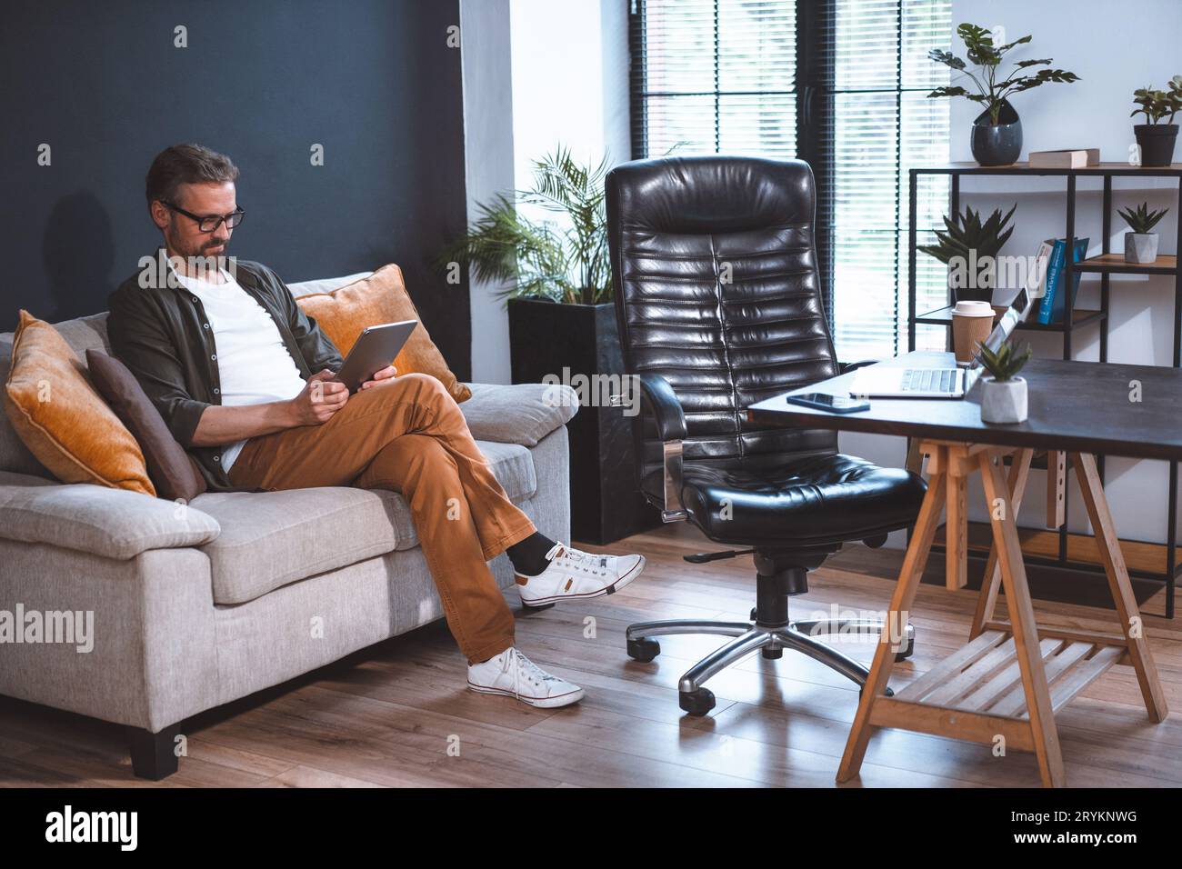 Freelancer sitting on sofa, looking relaxed as he uses tablet PC. Laptop on desk and empty chair can be seen nearby in room. Con Stock Photo