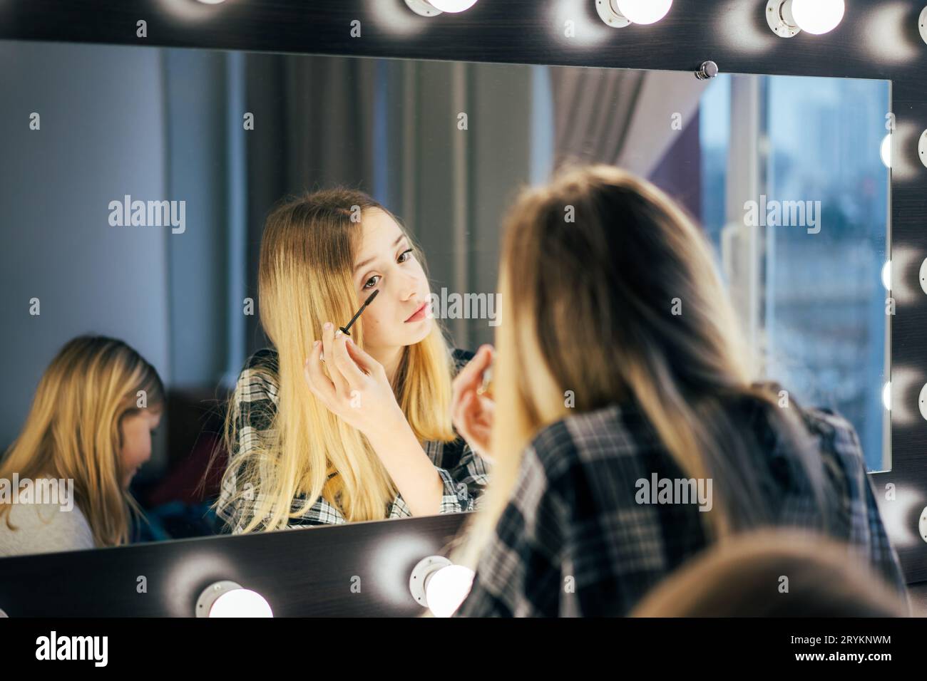 Young beautiful woman puts mascara on eyelashes in front of a large mirror in the dressing room. Stock Photo