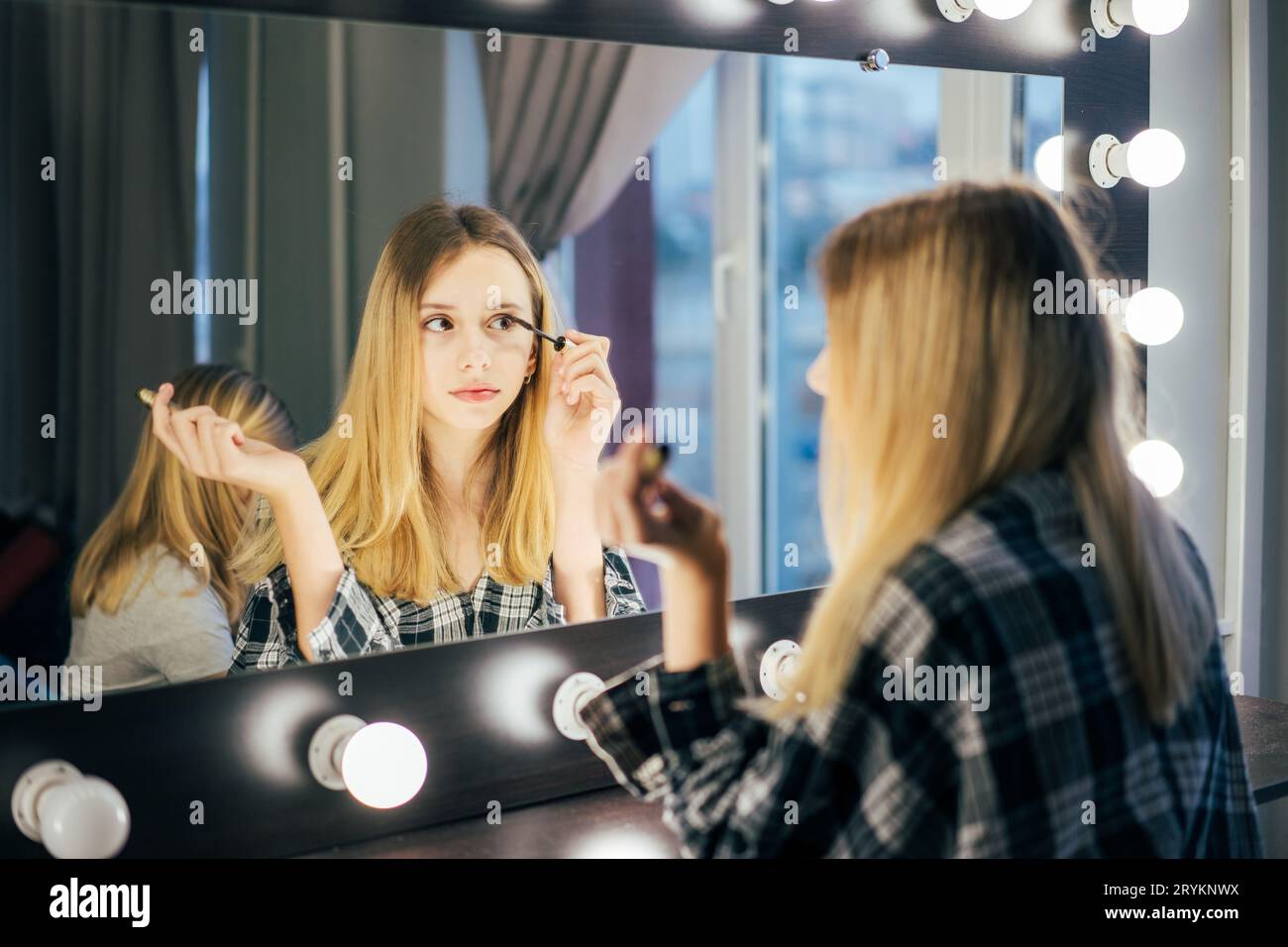 Stylish girl in puts mascara on the eyelashes. Teen in the dressing room in front of the mirror makes a make-up. person before t Stock Photo