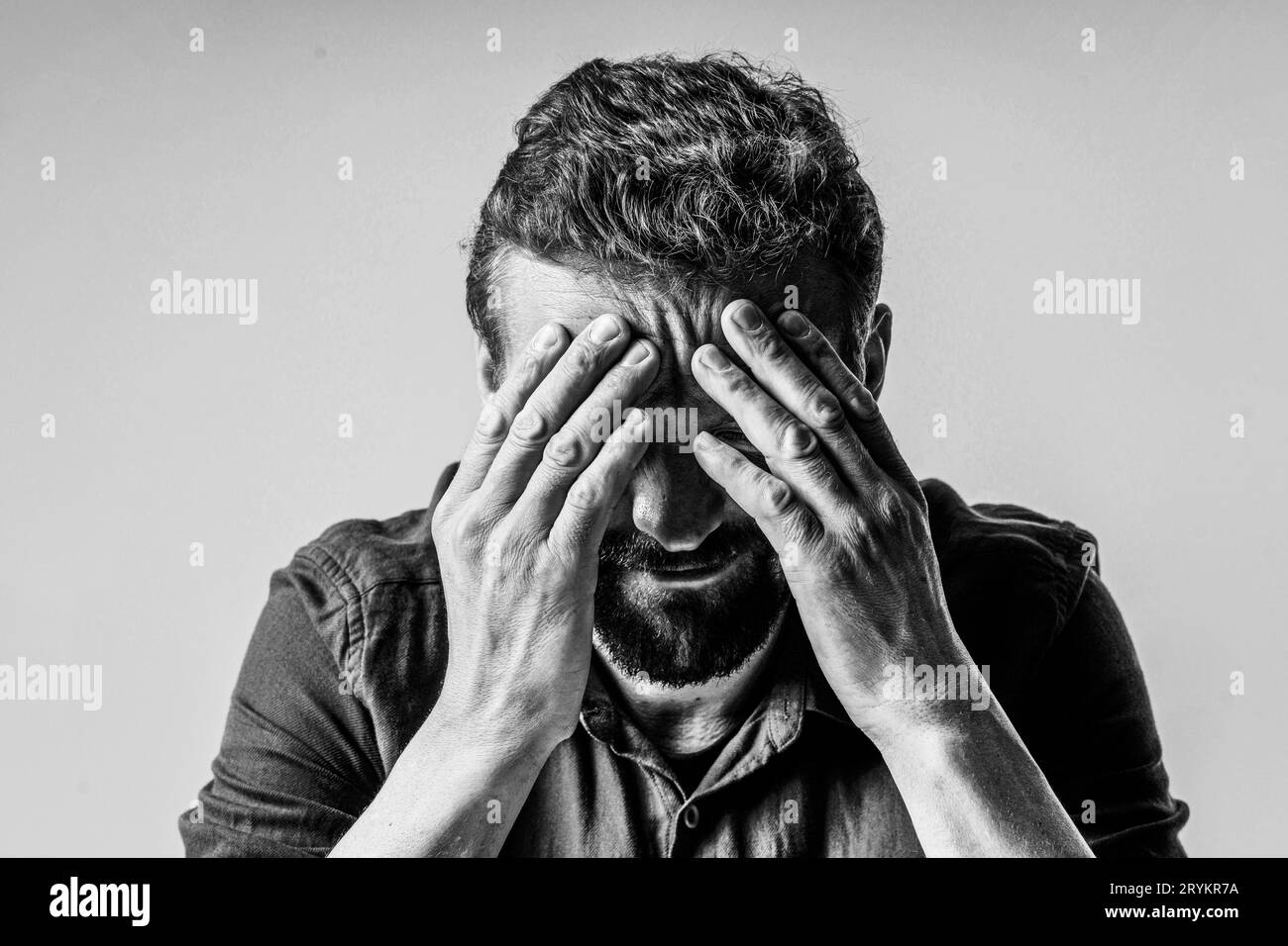 Black and white photo of a man looking extremely sad and depressed. He sitting on white background and has face covered with bot Stock Photo