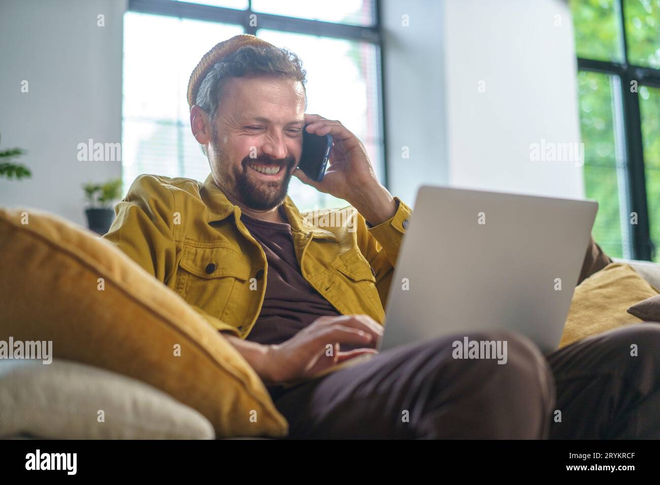 Smiling man having conversation on phone while working on laptop on sofa in office. He dressed in casual wear and most likely pr Stock Photo