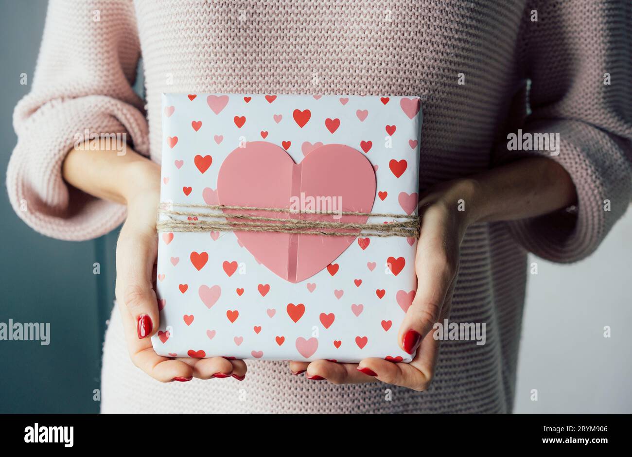 Wrapped gift for Valentine's Day decorated with a big pink heart in female hands. Stock Photo