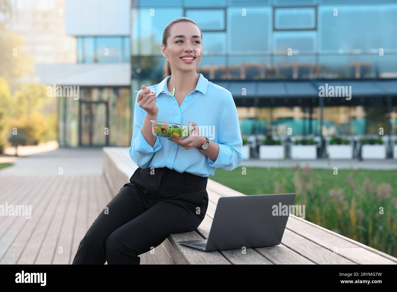 Smiling businesswoman with lunch box sitting near laptop outdoors Stock