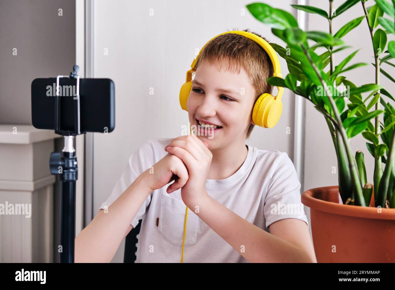 Boy blogger in yellow headphones talking with followers. Teenager live streaming looking to smartphone screen and gestures at ho Stock Photo