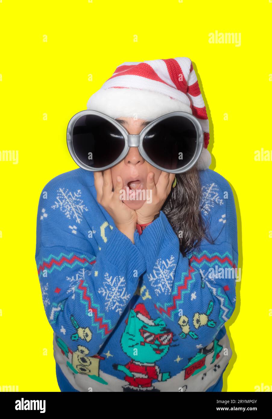 Woman with long hair, makes a surprised gesture to the camera. The woman is dressed in a blue ugly sweater, a Christmas hat, and large glasses. Studio Stock Photo