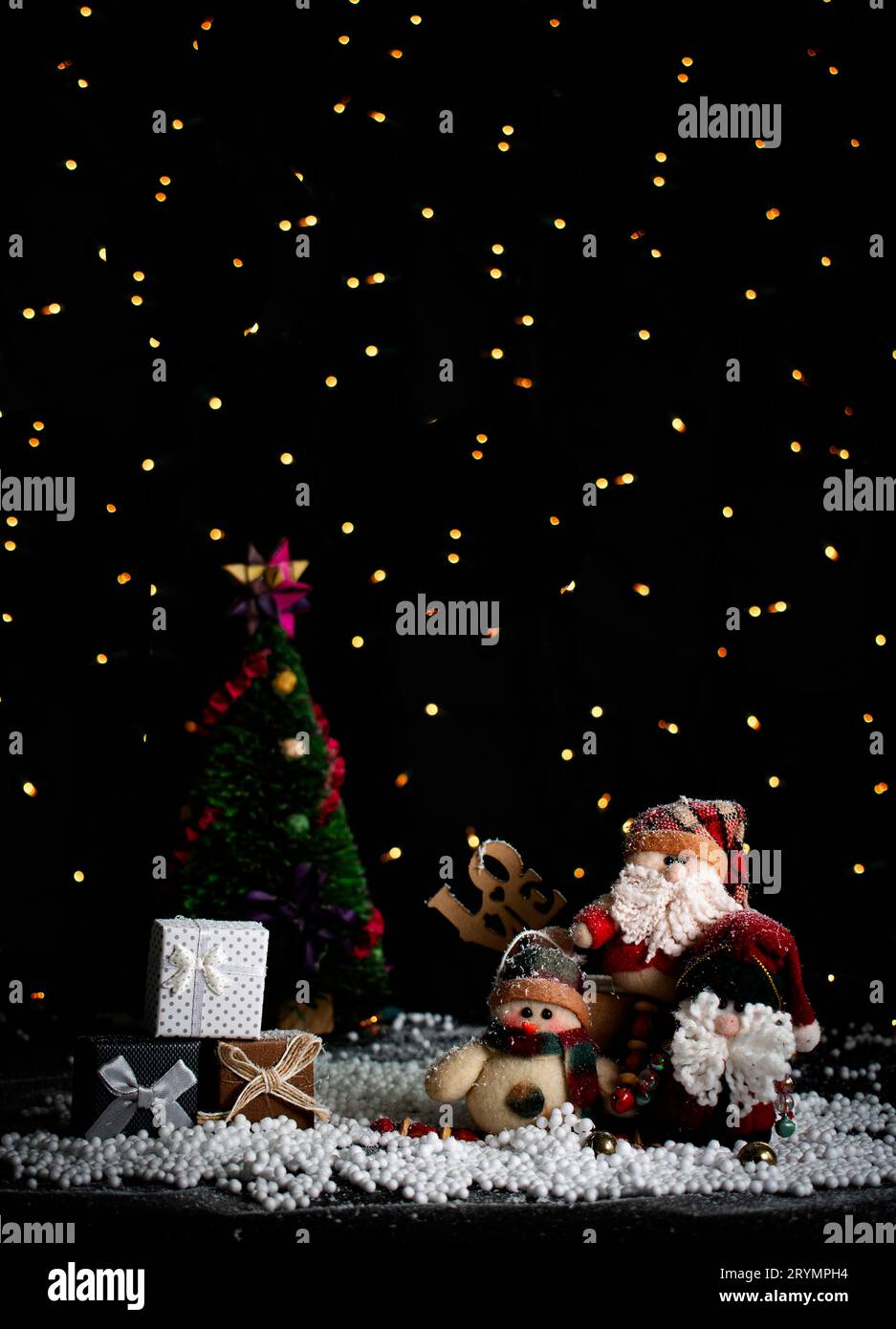 Christmas card featuring 3 plush Santas, 3 gift boxes, and in the background, a Christmas tree, all set against a black backdrop with blurred Christma Stock Photo