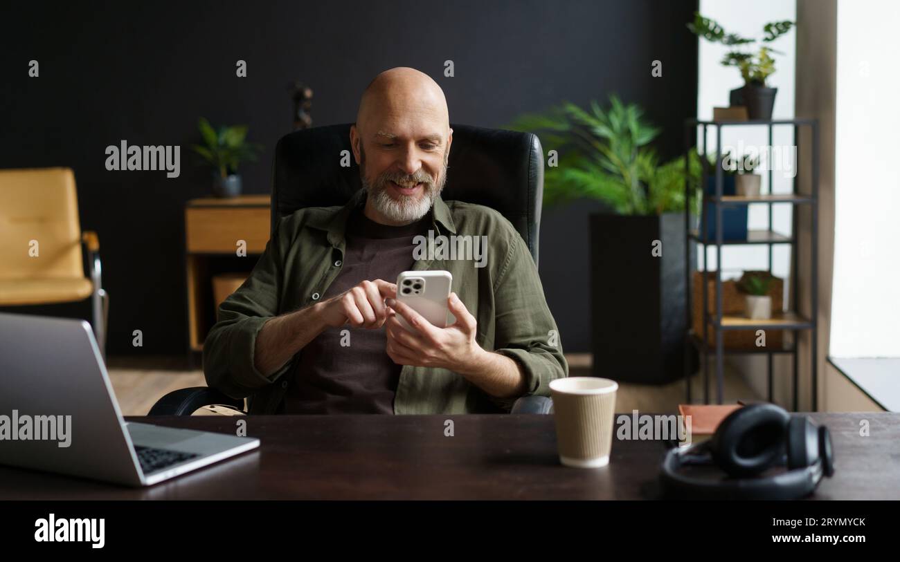 Bald and respectable man with gray beard. He captured in moment of joy, smiles while reading messages on phone. Computer, cup of Stock Photo