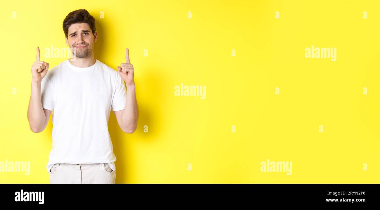 Unamused handsome guy frowning, pointing fingers up at something bad, standing skeptical against yellow background Stock Photo