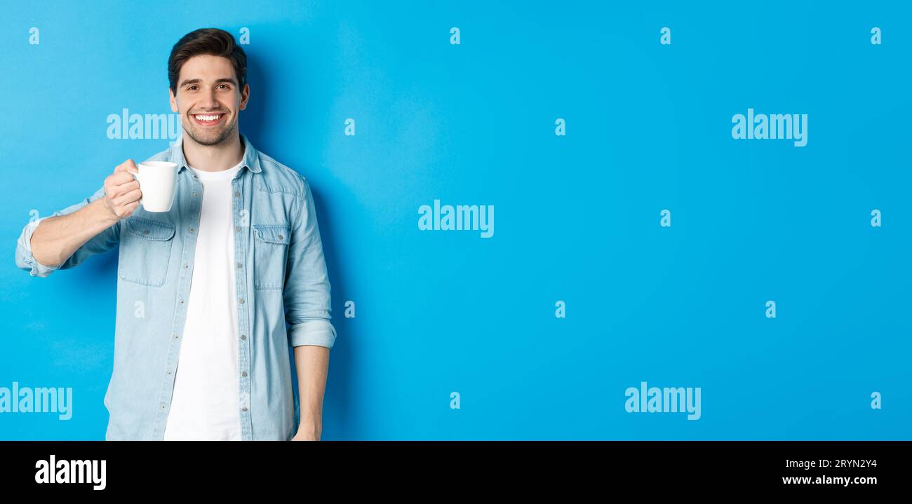 Smiling bearded man holding mug and drinking coffee, standing against blue background Stock Photo