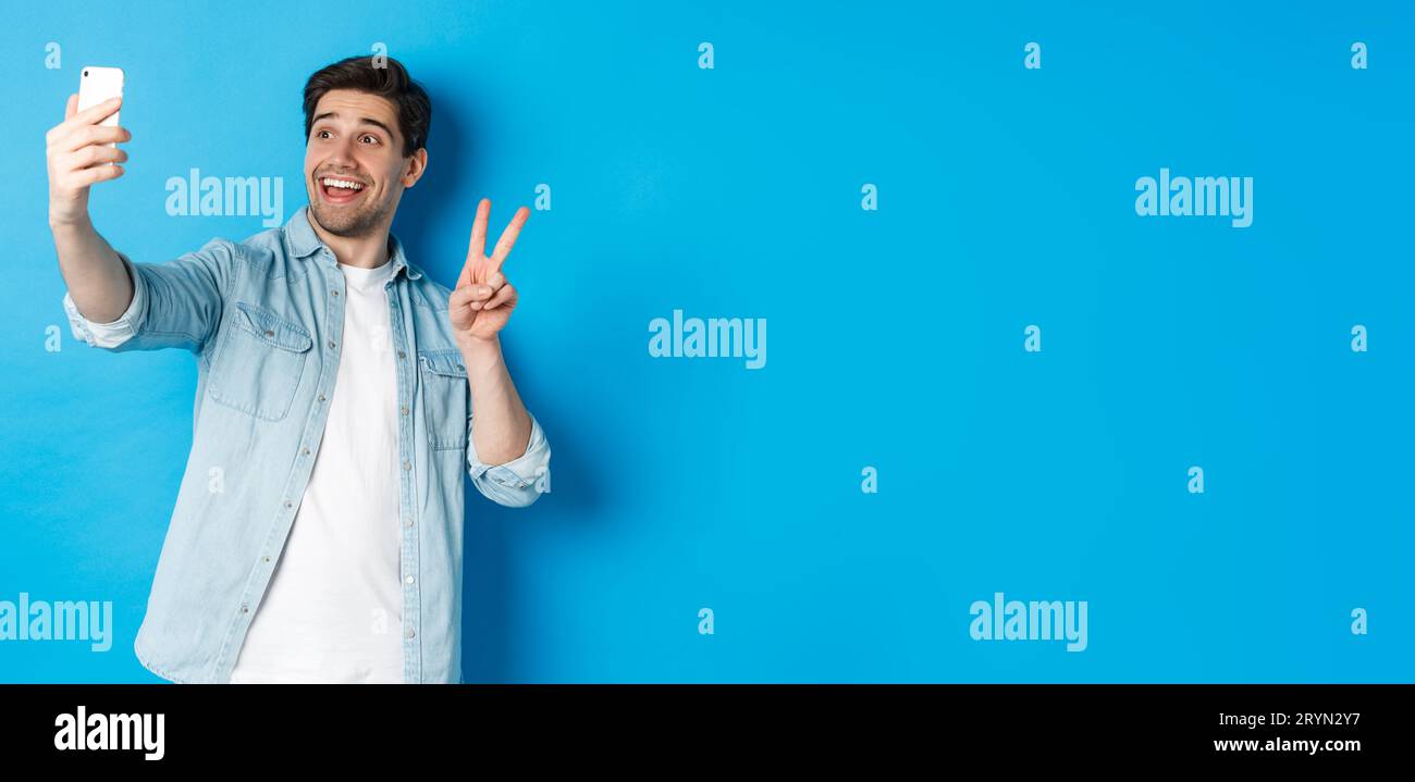 Happy man taking selfie and showing peace sign on blue background, holding mobile phone Stock Photo