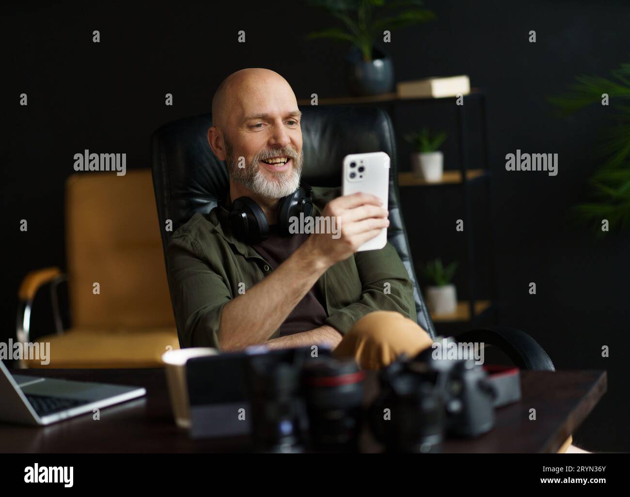 Mid aged man with bald head and silver beard engrossed in reading news on mobile phone. With laptop placed on desk in background Stock Photo