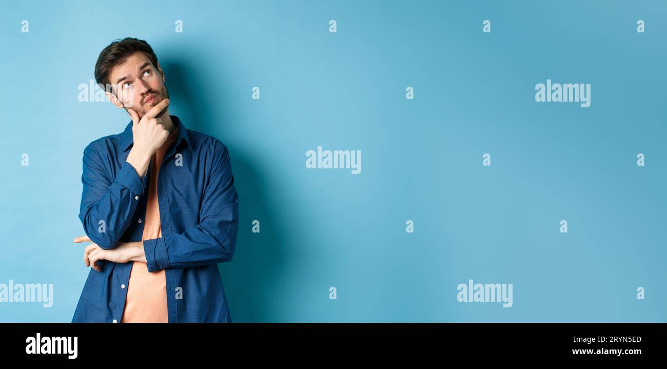 Pensive young man touching face and looking aside at empty space, imaging something, thinking off plan, standing on blue backgro Stock Photo