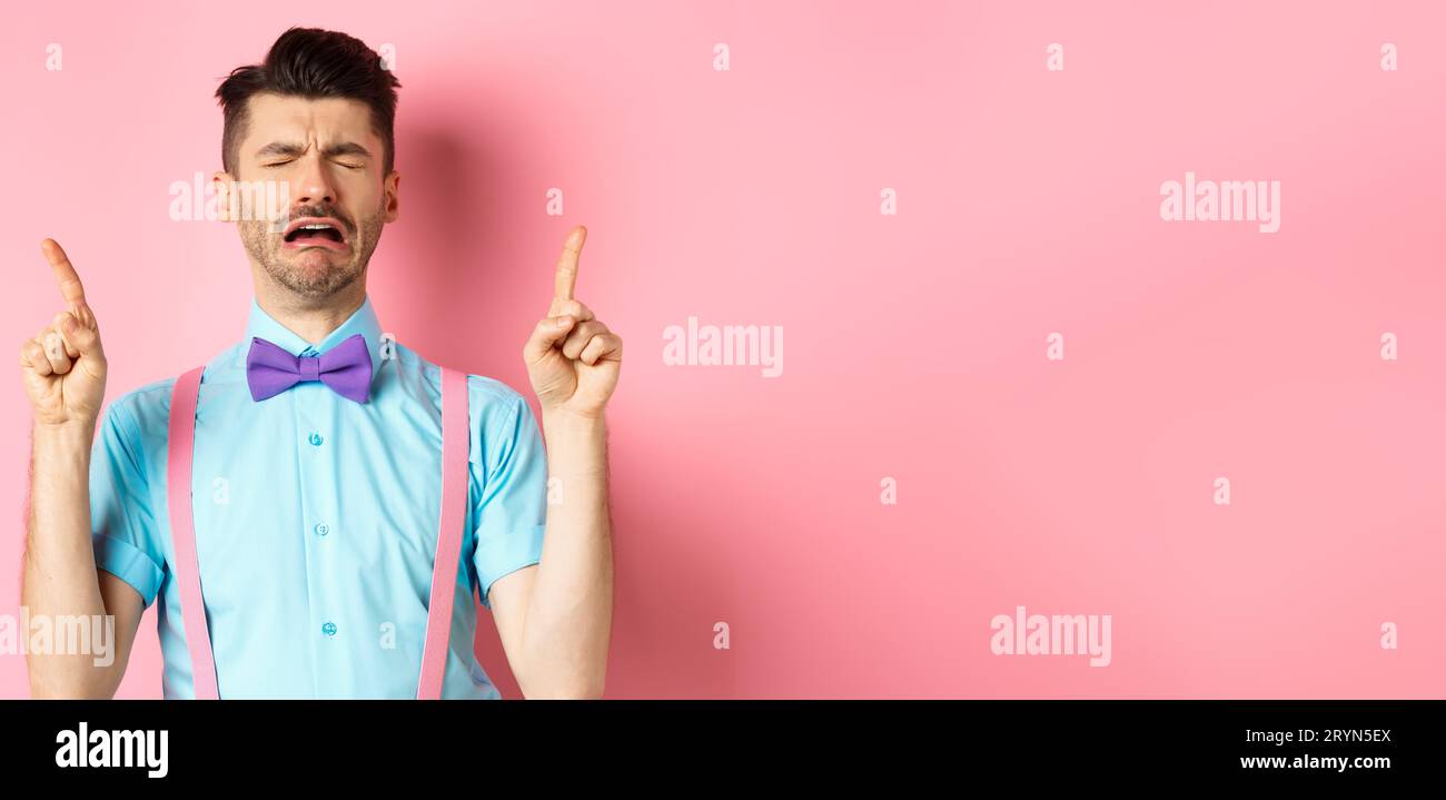 Sad and miserable guy sobbing and crying, pointing fingers up and something disappointed, standing upset on pink background Stock Photo