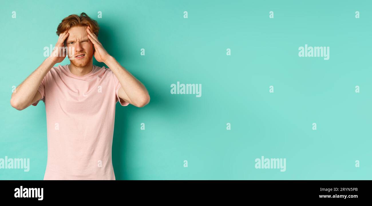 Young redhead man touching head and looking dizzy, feeling headache or migraine, standing in t-shirt over mint background Stock Photo