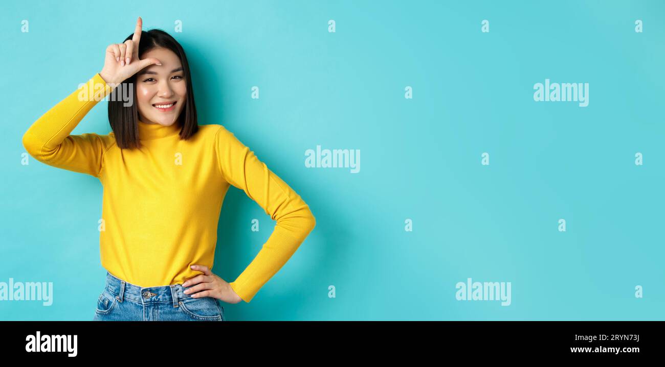 Sassy asian girl mocking lost team, showing loser sign on forehead and smiling pleased, being a winner, standing over blue backg Stock Photo