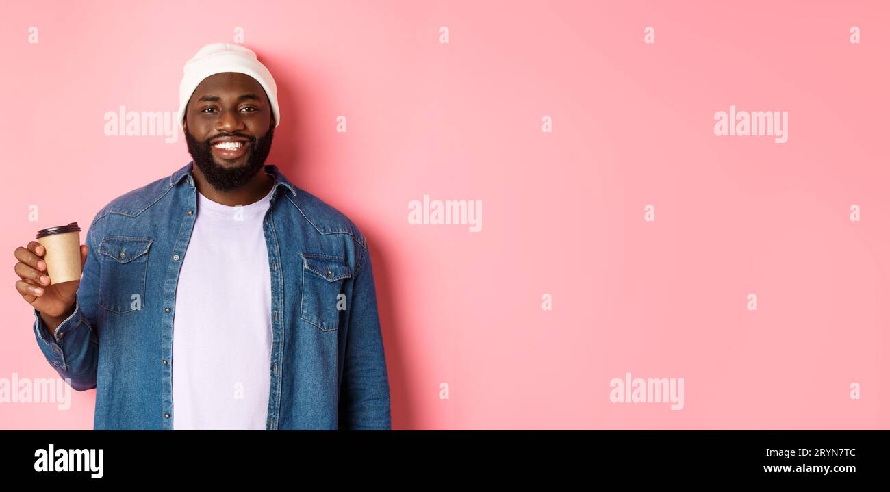 Handsome modern Black man drinking takeaway coffee, smiling and looking satisfied at camera, standing over pink background Stock Photo