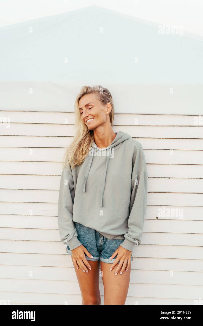 Cute beautiful tanned blonde woman in denim shorts and a stylish hoodie having fun  and sincerely laughs. Stock Photo