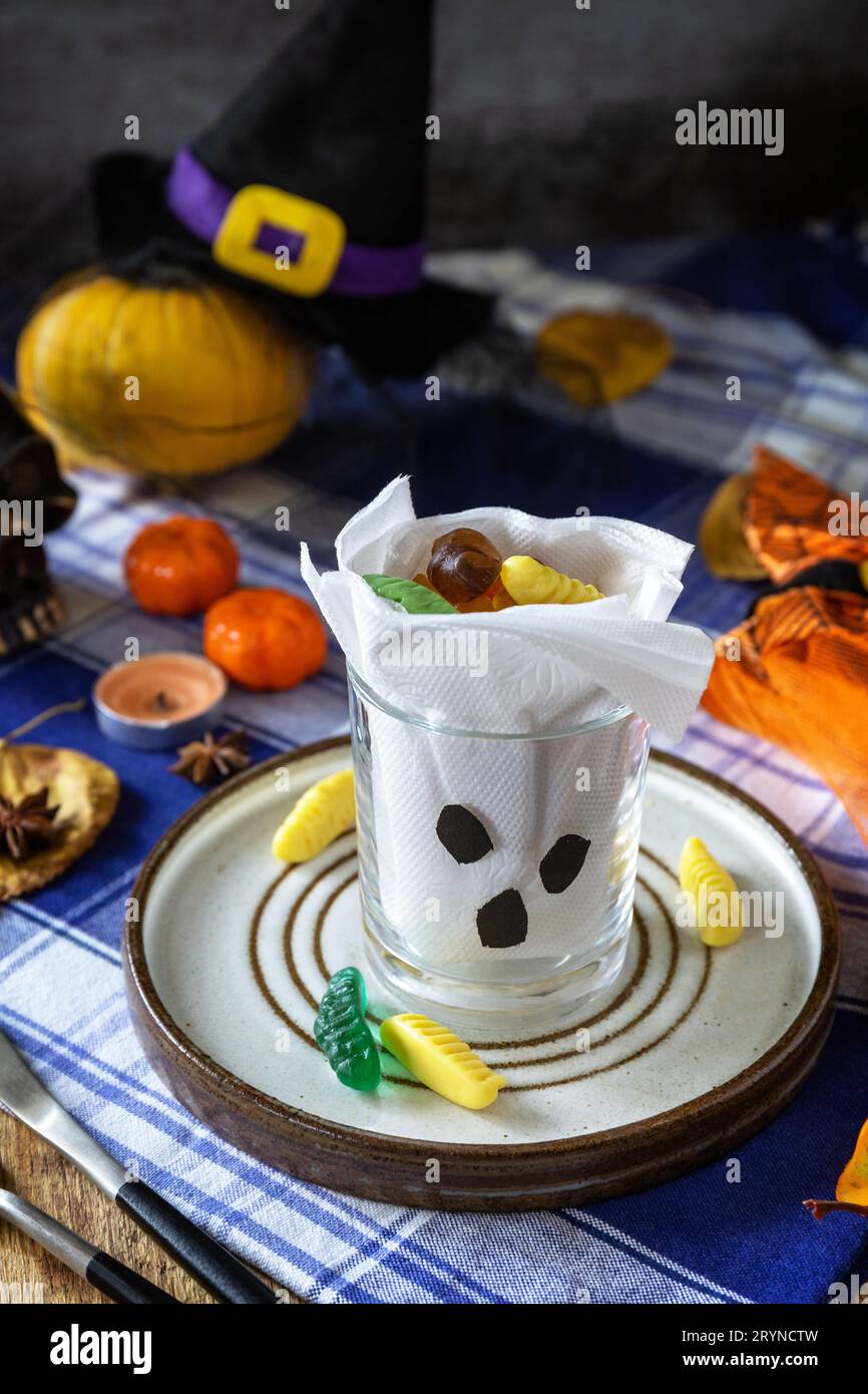 Halloween party. White ghost crafts scary face in glass on a plate and a festive Halloween table setting with pumpkins. Stock Photo