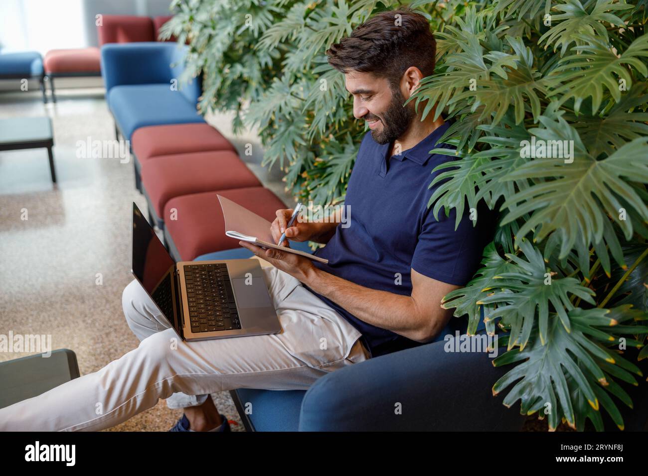 Handsome smiling freelancer is making notes during work on laptop in office hall Stock Photo