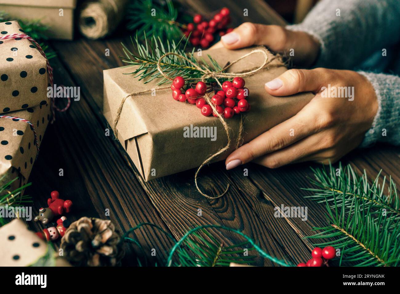 Close-up of a Christmas present wrapped in craft paper and decorated with berries and a spruce branch in female hands. Stock Photo
