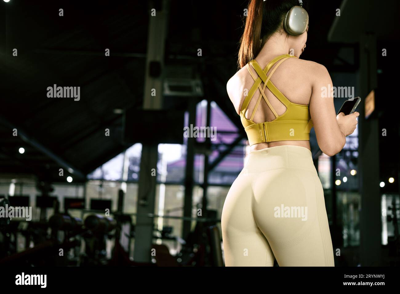 Young asian woman wearing headphones listening to music from her smart phone. Female fitness model in gym using mobile phone. Stock Photo