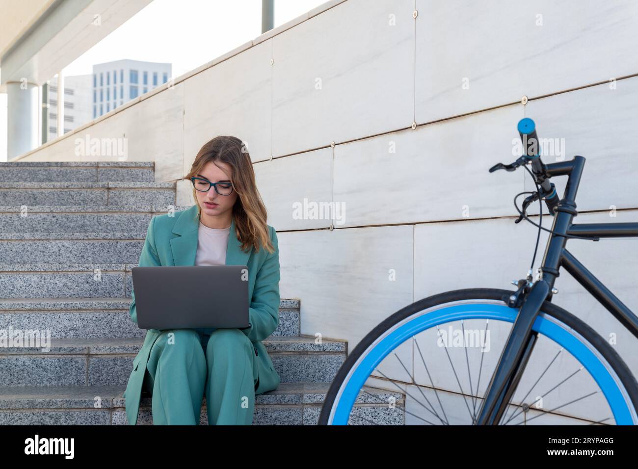 Business woman sitting on stairs outside office building while working with laptop next to a bike Stock Photo
