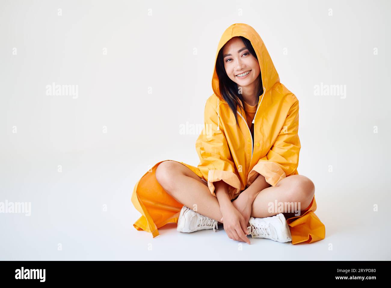 Smiling asian girl with braces dressed in bright yellow raincoat posing with hood on her head on white studio background sitting on floor with copy sp Stock Photo