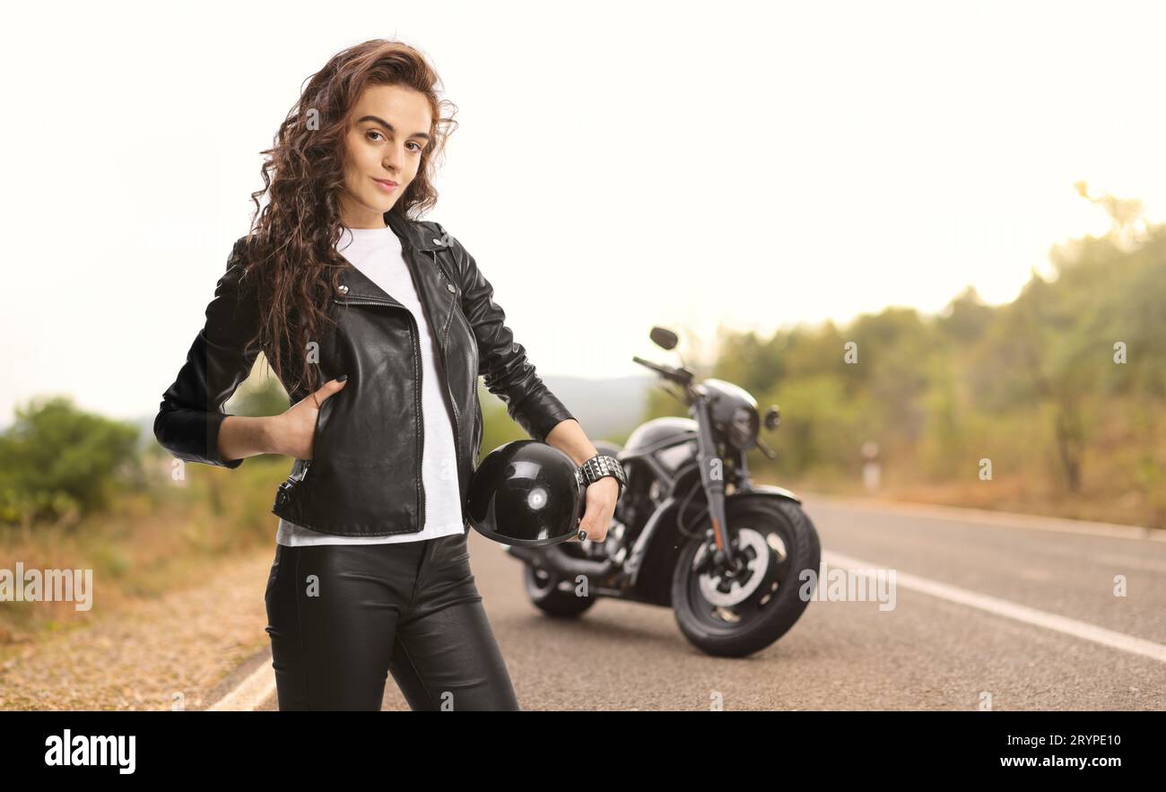 Female biker in a leather jacket holding a helmet and posing on the road Stock Photo
