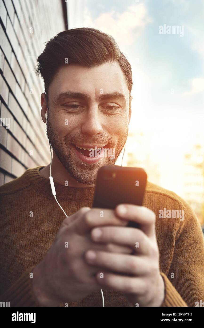 Portrait of smiling man with headphones and cellphone standing by brick wall. Stock Photo