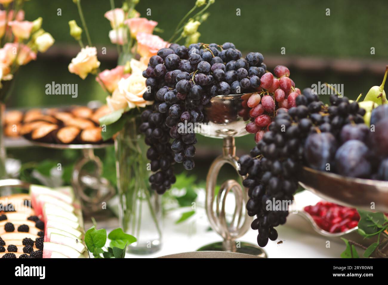An array of silver dishes filled with purple grapes, each one topped with a colorful vase filled with blooming flowers Stock Photo